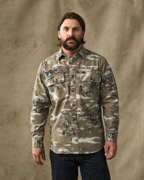A man wearing a long-sleeve button-up shirt with a detailed landscape print in beige, brown, and gray tones, paired with dark blue jeans, standing against a textured beige backdrop.