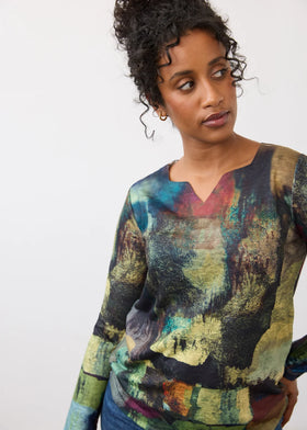 A woman wearing a long-sleeve V-neck top with an abstract, painterly multicolor print in shades of green, yellow, blue, and brown, set against a plain light gray background.
