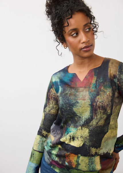 A woman wearing a long-sleeve V-neck top with an abstract, painterly multicolor print in shades of green, yellow, blue, and brown, set against a plain light gray background.