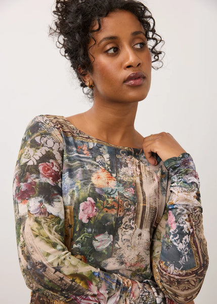 A woman wearing a long-sleeve boat-neck top with a complex, painterly floral and botanical print in muted tones of green, beige, pink, and blue, set against a plain light gray background.