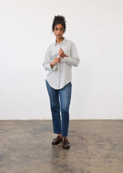 A woman stands in a minimalist studio setting with a white wall and concrete floor, wearing a white button-down shirt with thin black vertical stripes, blue jeans, and dark brown leather loafers.