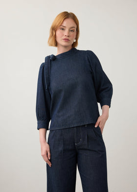 A woman modeling a dark indigo blue long-sleeve top with a unique asymmetrical tie detail at the collar, paired with matching dark denim pants, against a plain light gray studio background.