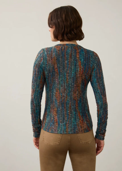 A woman viewed from the back wearing a long-sleeve knit top with a vertical marled pattern in teal, blue, rust orange, and brown, paired with tan pants, against a plain light gray background.