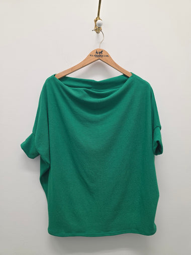 A green short-sleeve top with a draped, wide neckline hanging on a wooden hanger against a plain white wall.