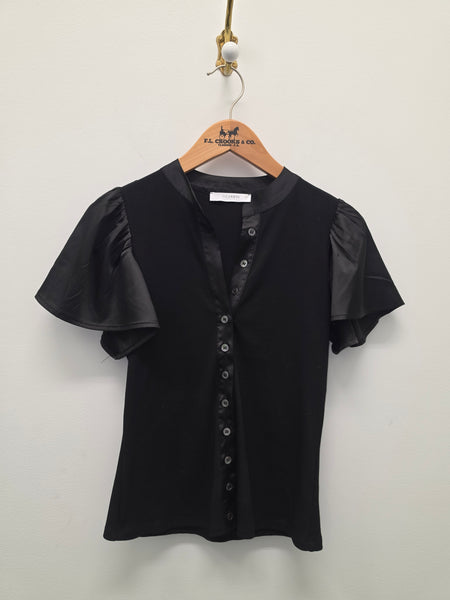 A black short-sleeve blouse with sheer flutter sleeves and a button-front neckline, hanging on a wooden hanger against a plain light wall.
