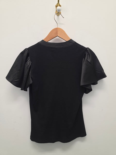 A black short-sleeve top with textured, ruffled flutter sleeves, hanging on a wooden hanger against a plain off-white wall.