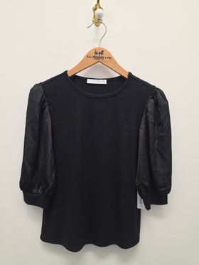 A black short-sleeve top with sheer puff sleeves hanging on a wooden hanger against a plain off-white wall.