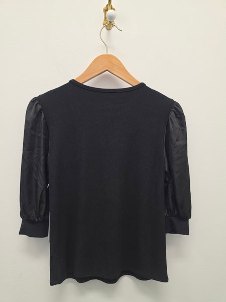 A black short-sleeve top with sheer puff sleeves hanging on a wooden hanger against a plain off-white wall.