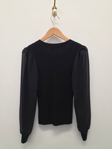 A black long-sleeve top with sheer puff sleeves hanging on a wooden hanger against a plain off-white wall.