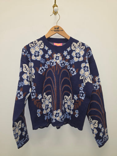 A navy blue knit sweater with a white and light blue floral pattern and brown central detailing, hanging on a wooden hanger with the brand name 'S.A.