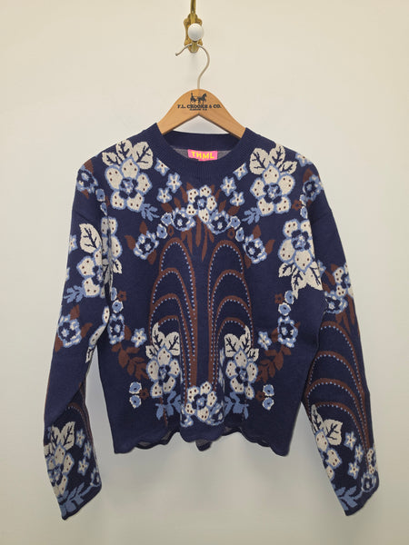 A navy blue knit sweater with a white and light blue floral pattern and brown central detailing, hanging on a wooden hanger with the brand name 'S.A.