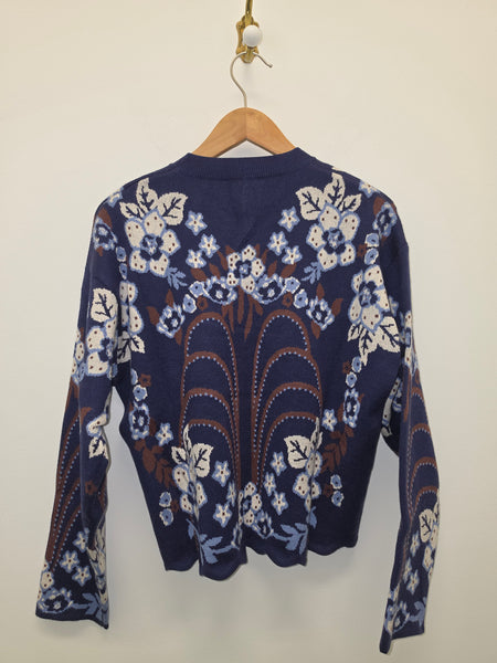 A navy blue knit sweater with intricate floral and leaf patterns in cream, light blue, and brown, hanging on a wooden hanger against a plain off-white wall.