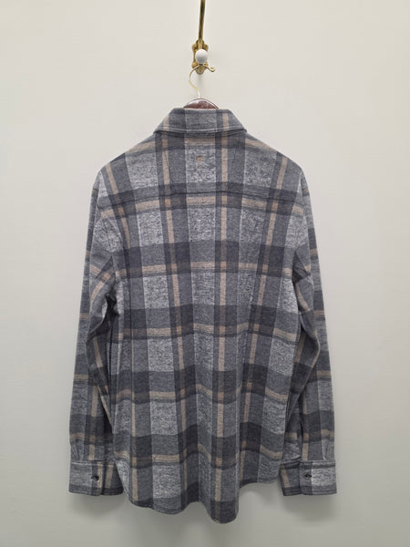 A long-sleeve plaid flannel shirt in gray, black, and beige, hanging on a hanger against a plain off-white wall.