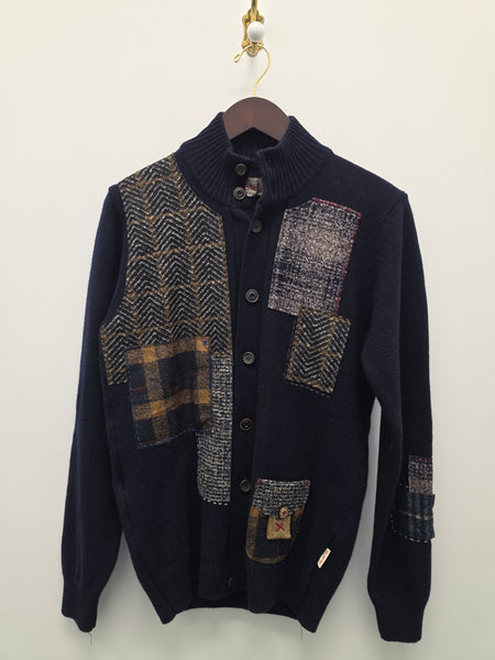 A dark navy blue knit cardigan sweater with patchwork herringbone and plaid fabric details, shown hanging on a hanger against a plain off-white wall.