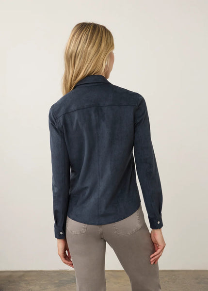 A woman viewed from behind wearing a dark navy blue long-sleeve shirt and gray pants, standing against a plain off-white wall.