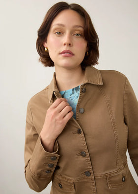 A woman wearing a tan utility jacket over a blue patterned top, photographed against a plain light gray background.