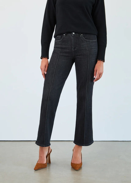 A model from the waist down wearing dark charcoal gray straight-leg jeans with contrasting light stitching, paired with a black sweater and brown pointed-toe heels against a light gray studio background.