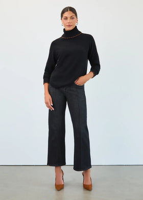 A woman modeling a black turtleneck sweater with gold chain detailing at the collar, paired with dark wide-leg jeans and brown pointed-toe heels, standing against a plain white studio wall.