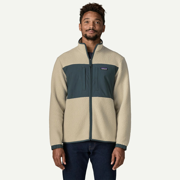 A man wearing a Patagonia fleece jacket in beige and dark teal, paired with dark blue jeans, standing against a light off-white background.