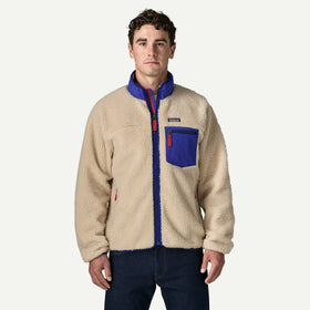 A man wearing a Patagonia fleece jacket in beige with royal blue collar, zipper placket, and chest pocket, paired with dark blue jeans, against a light gray studio background.