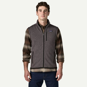 A man wearing a Patagonia fleece vest with brown and black plaid sleeves, layered over a collared shirt and paired with dark blue jeans, against a light gray background.