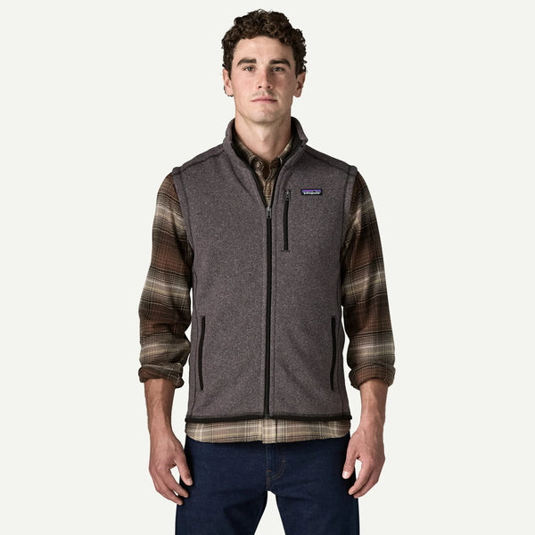 A man wearing a Patagonia fleece vest with brown and black plaid sleeves, layered over a collared shirt and paired with dark blue jeans, against a light gray background.