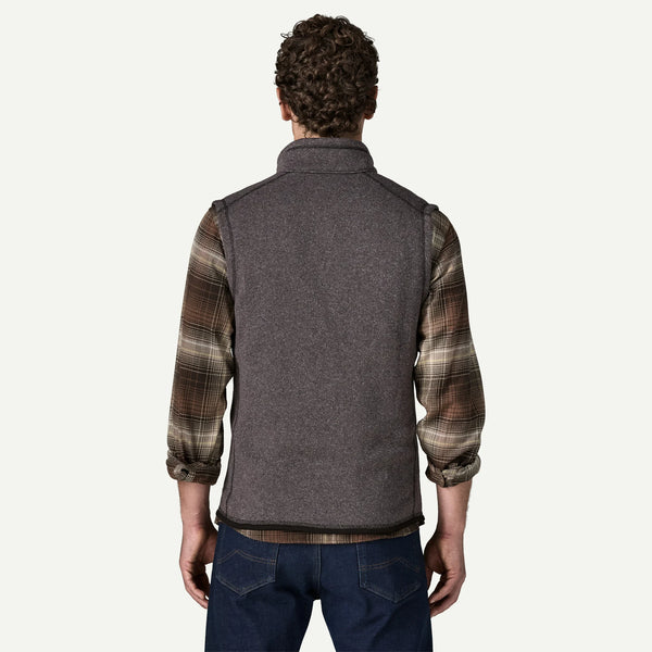 A man viewed from the back wearing a dark gray fleece vest over a brown and beige plaid flannel shirt, paired with dark blue jeans, against a light off-white background.
