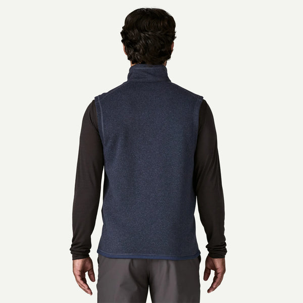 A man viewed from the back wearing a navy blue sleeveless quarter-zip fleece vest over a black long-sleeve shirt and gray pants, against a light off-white background.