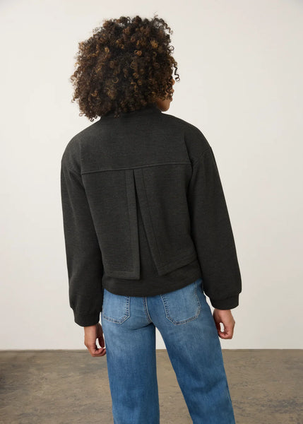 A person shown from the back wearing a dark charcoal gray cropped jacket with a unique pleated back detail and blue jeans, standing against a plain off-white wall.
