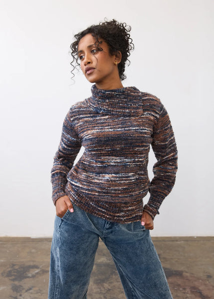A woman wearing a marled, textured knit mock neck sweater in a mix of brown, blue, and cream tones, paired with wide-leg blue jeans, standing against a plain white wall.