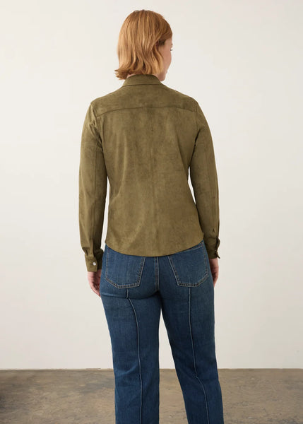 A woman viewed from behind wearing an olive green long-sleeve shirt and blue jeans, standing against a plain off-white wall.
