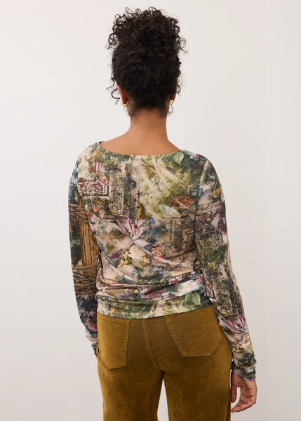 A woman viewed from behind wearing a long-sleeve top with a detailed multicolor botanical and architectural print, paired with brown pants, against a plain off-white background.