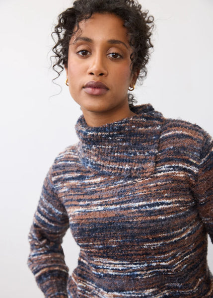 A woman wearing a marled, textured knit mock neck sweater with horizontal stripes of navy, brown, beige, and white, photographed against a plain white background.