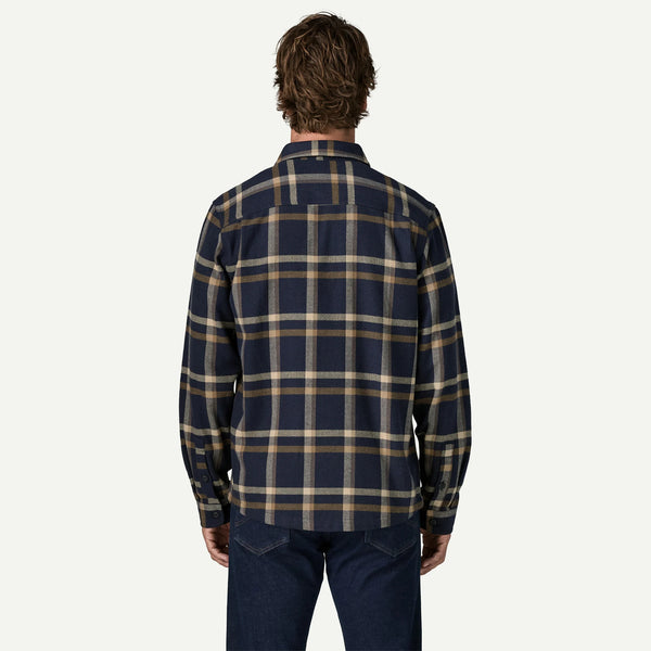A man viewed from behind wearing a navy blue and beige plaid button-down shirt with dark blue jeans, set against a plain off-white background.