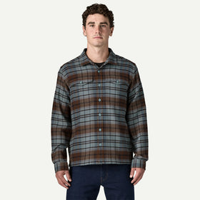 A man wearing a long-sleeve button-up plaid flannel shirt in brown, gray, and black, paired with dark blue jeans, against a light off-white background.
