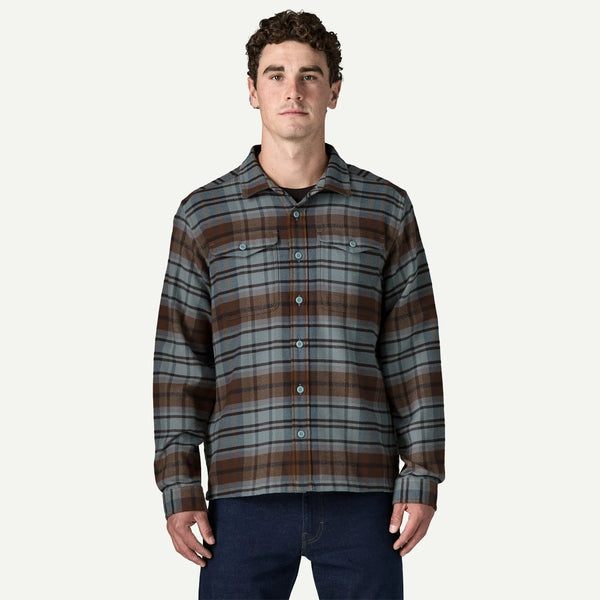 A man wearing a long-sleeve button-up plaid flannel shirt in brown, gray, and black, paired with dark blue jeans, against a light off-white background.