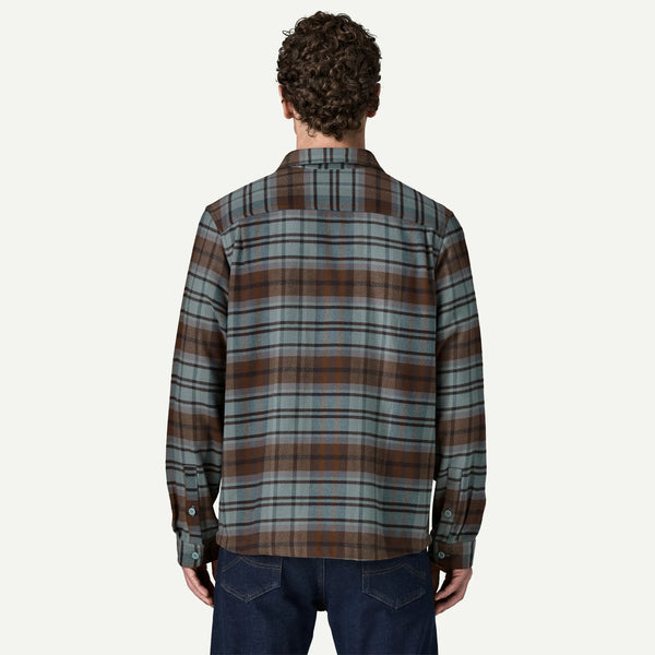 A man viewed from behind wearing a long-sleeve plaid button-up shirt in muted green, brown, and gray tones, paired with dark blue jeans, against a solid off-white background.