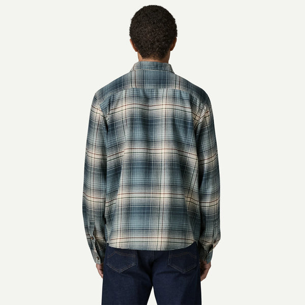 A person viewed from the back wearing a long-sleeve plaid button-up shirt in muted blue, beige, and gray tones, paired with dark blue jeans, against a plain light background.