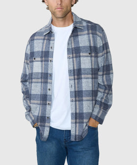 A man wearing a blue and gray plaid button-down shirt over a white t-shirt and blue jeans, shown from the waist up against a light gray background.