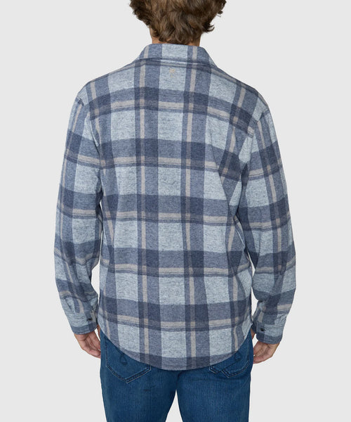 A man viewed from behind wearing a long-sleeve plaid flannel shirt in shades of blue, gray, and beige, paired with blue jeans, against a light gray studio background.