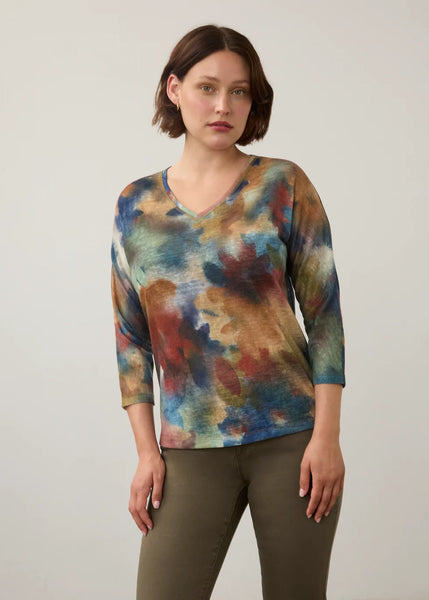 A woman modeling a three-quarter sleeve V-neck top with an abstract watercolor tie-dye pattern in blue, brown, red, and green, paired with olive green pants, against a plain light gray background.