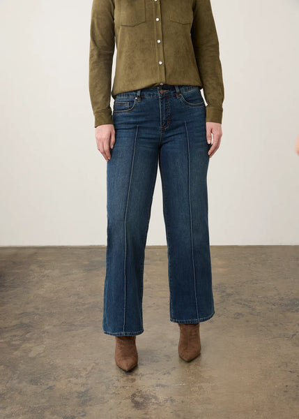 A model wearing a pair of dark blue wide-leg cropped jeans, paired with an olive green button-up shirt and brown suede boots, standing against a neutral studio backdrop.
