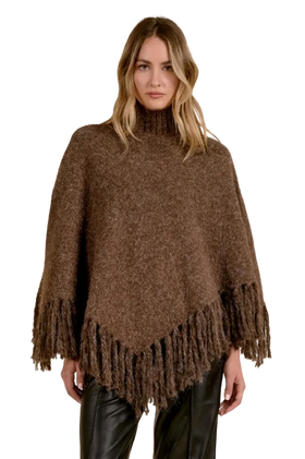 A woman modeling a brown fuzzy knit poncho with fringe trim, paired with black leather pants, against a plain white background.