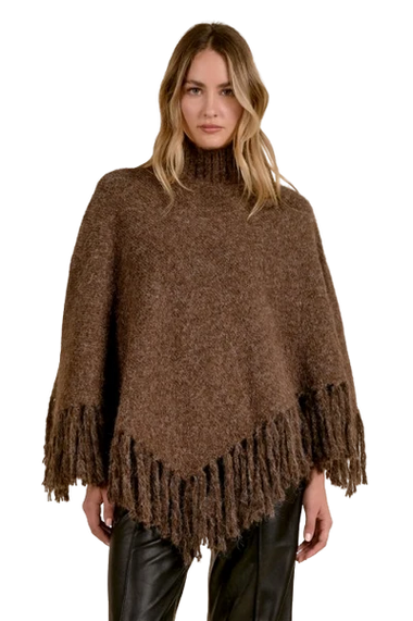 A woman modeling a brown fuzzy knit poncho with fringe trim, paired with black leather pants, against a plain white background.