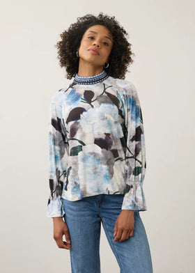 A woman with curly dark hair wearing a long-sleeve blouse with a blue, white, and black abstract floral print, paired with blue jeans, standing against a plain light background.