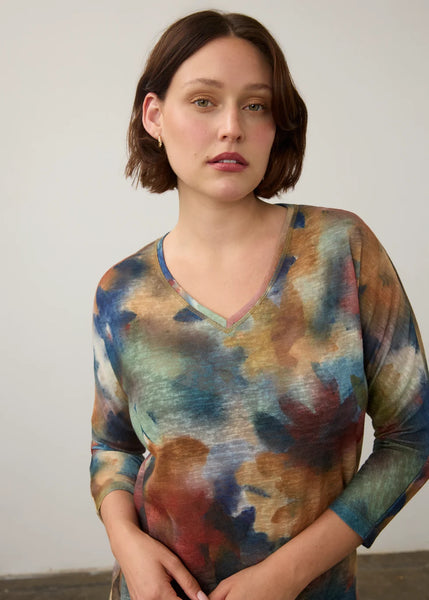 A woman with short brown hair wearing a V-neck long-sleeve top with an abstract watercolor print in shades of blue, green, brown, and beige, standing against a plain light gray wall.