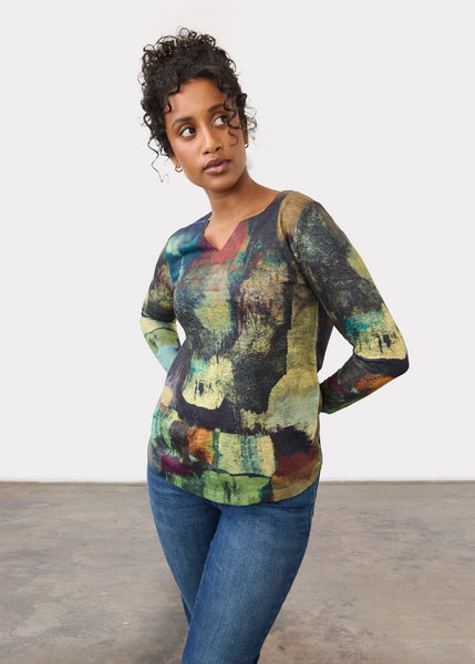 A woman modeling a long-sleeve V-neck top with an abstract multicolor print in shades of green, yellow, brown, and blue, paired with blue jeans against a plain white wall.