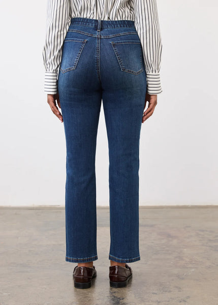 Rear view of a person wearing high-waisted blue jeans, a striped long-sleeve shirt, and brown leather loafers against a plain indoor wall.
