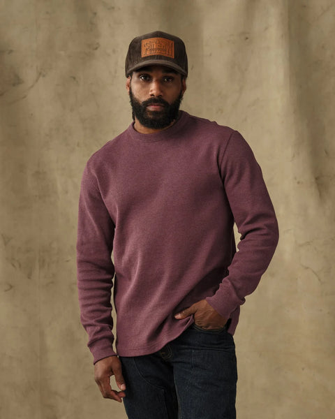 A man wearing a heathered dusty rose long-sleeve crewneck sweater, dark blue jeans, and a brown trucker hat with a tan leather patch, standing against a textured beige studio backdrop.