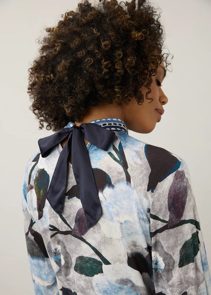 A close-up back view of a person with curly dark hair wearing a long-sleeve blouse with a large bow at the back and an abstract blue, white, and gray pattern.